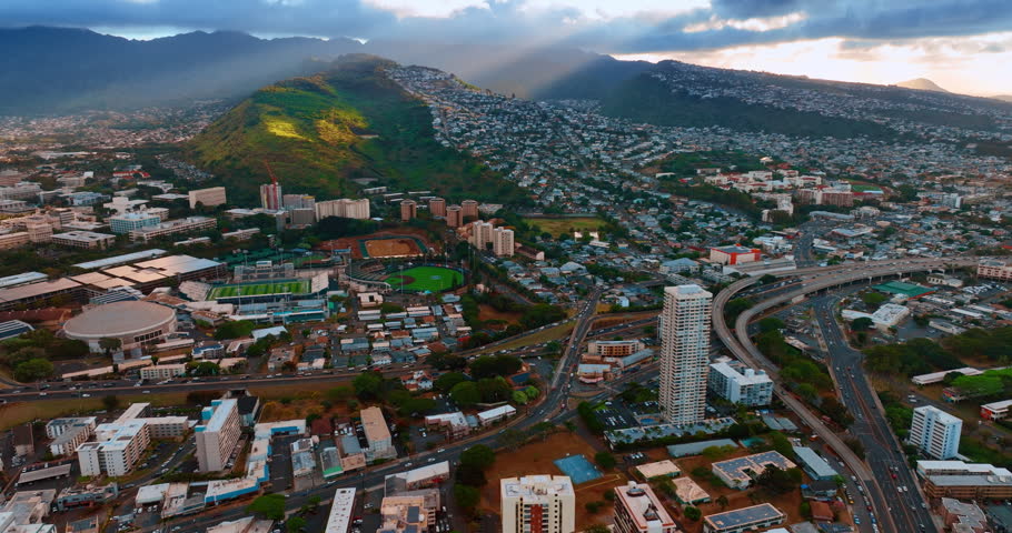 System of roads crossing the cityscape of Honolulu, Hawaii, USA. Multiple cars move by the highway. Sunrays light the verdant mountains. Aerial view.