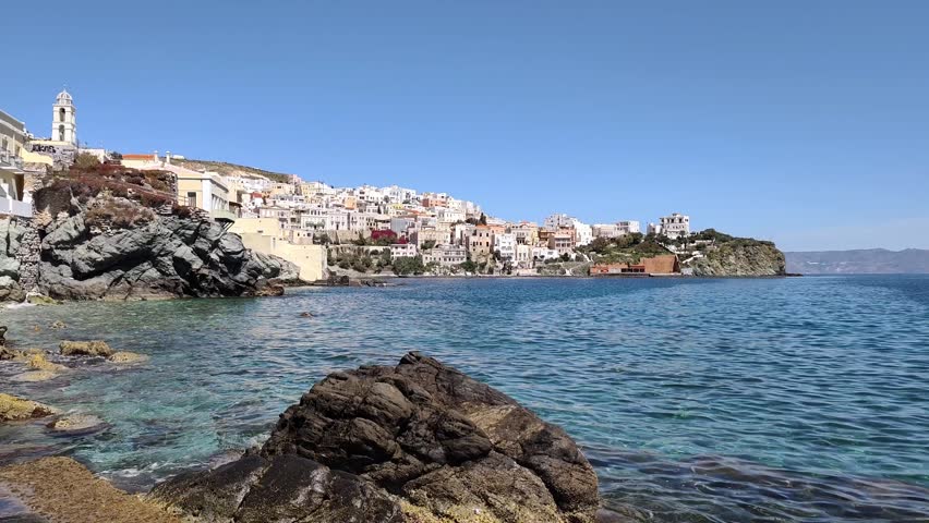 View of the picturesque Vaporia district and Agios Nikolaos Church viewed from Kymata, a coastal quarter of Ermoupoli on Syros island, Greece, with elegant neoclassical mansions cascading toward the turquoise, crystal-clear waters of the Aegean Sea beneath a bright blue sky.