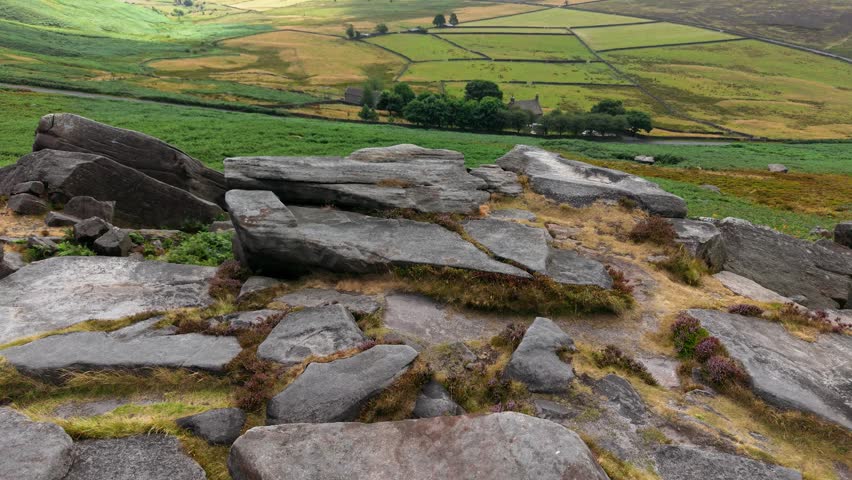 Rocky outcrop overlooking green patchwork farmland and rural countryside. Weathered stone slabs with moorland vegetation above fields and country landscape. Scenic geology with natural terrain for