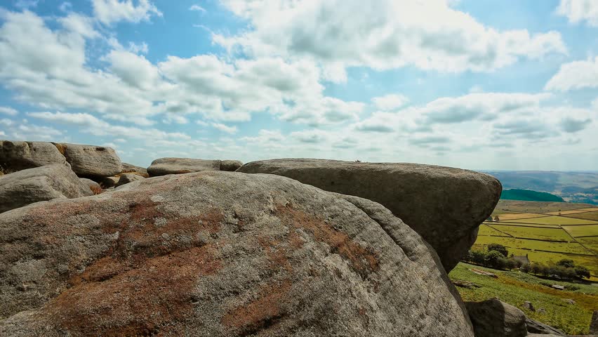 Rounded granite boulders with expansive countryside panorama slide shot. Weathered rock surface beside agricultural fields and layered ridgelines. Open moorland landscape beneath scattered white