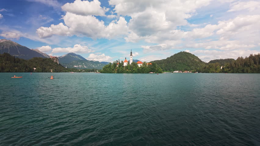Low-angle view across the surface of Lake Bled in Slovenia toward Bled Island and the iconic Church of the Assumption, with boats, paddleboarders and the Julian Alps on a sunny day.