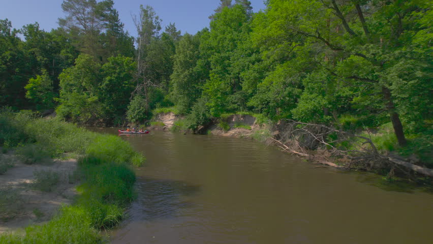Friends tandem kayaking on a broad forest river features two inflatable boats paddling separately through a gentle bend while a low lateral drone follows beside grass eroded banks and dense trees