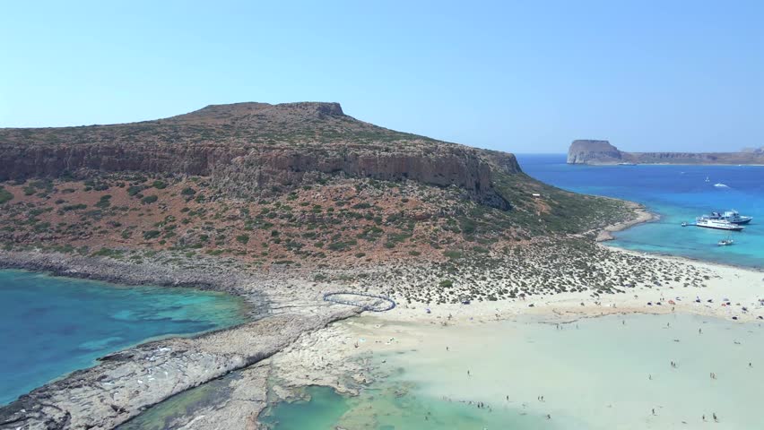 Beautiful wide drone shot panning right over the exotic turquoise waters and white sand of Balos lagoon in Crete, Greece. Features anchored tour boats and Gramvousa island on a sunny summer day.