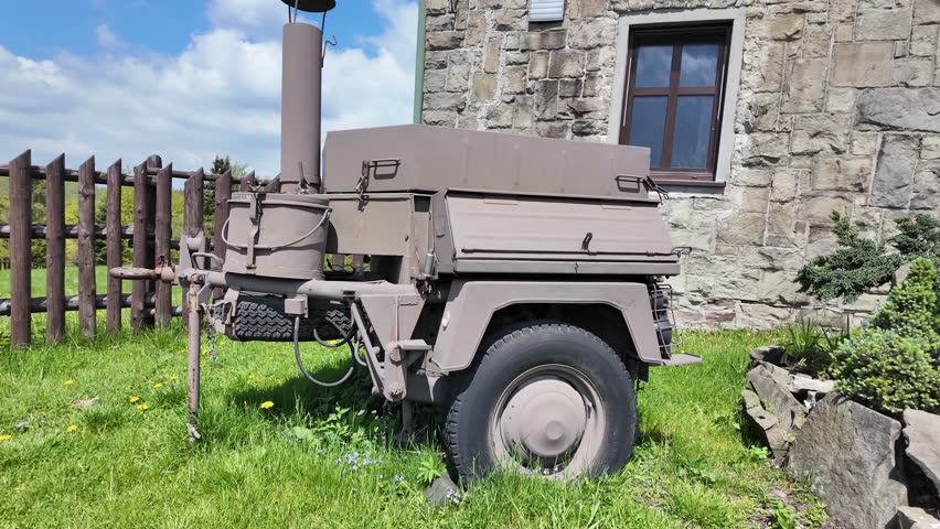 Restored portable field kitchen exhibited behind a wooden fence on green grass near a mountain lodge under bright summer sunshine and clear blue sky.
