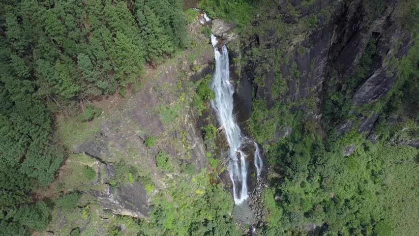 Scenic high aerial view of a dramatic waterfall plunging down a towering rocky cliff framed by pine tree forests and misty green mountains in the highlands of Sri Lanka on a cloudy day.