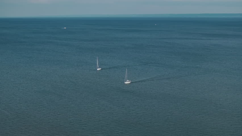 Two white sailboats navigate across the calm blue waters of Lake Ontario. Gentle ripples and light wakes stretch behind them under a hazy summer sky in Oakville, Ontario.