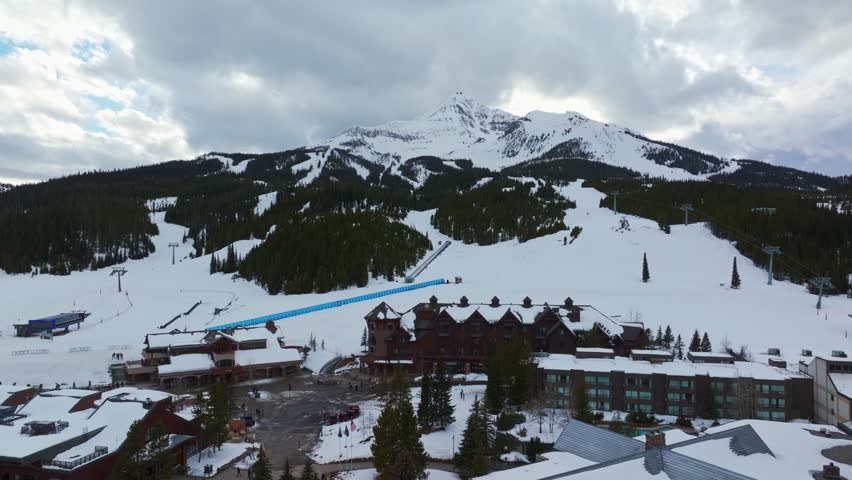 Trucking left aerial drone shot of a winter landscape of a mountain resort in Montana with Lone Peak rising in the background, surrounded by pine forests, homes, and snow-covered terrain.