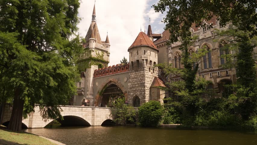 Static locked off view of Vajdahunyad Castle rising above the City Park lake and its stone bridge in Budapest, Hungary, on a sunny summer day.