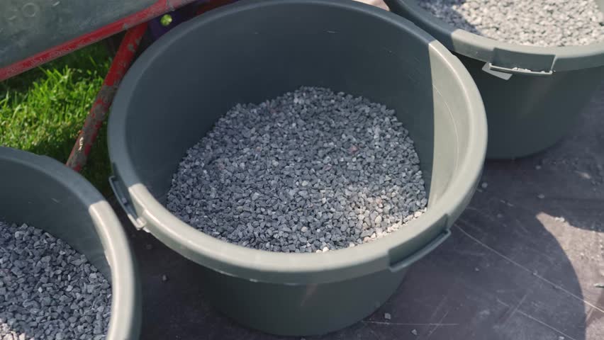 Closeup of gray gravel inside dark plastic buckets on paved ground. Bright daylight highlights texture shadows. Green grass red wheelbarrow in background