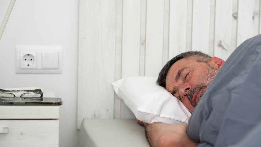 Mature man with a beard waking up in his bed, putting on his glasses and checking his mobile phone before going back to sleep