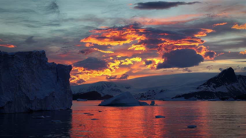 The vibrant orange and yellow hues of a sunset paint the sky above a serene Antarctic landscape with floating icebergs and majestic mountains reflected in the calm water
