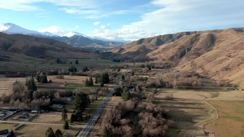 Aerial view of the scenic Burkes Pass landscape featuring rolling hills, snow-capped mountains, and a winding road in Burkes Pass, Canterbury Region, New Zealand.