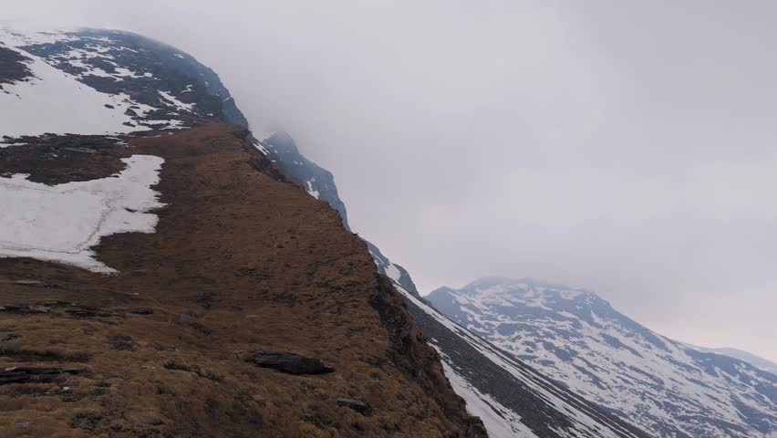 Drone aerial of a dangerous snow-covered mountain ridge hidden beneath dense fog in the Himalayas. Perfect for adventure documentaries, survival films, and cinematic mountain visuals.