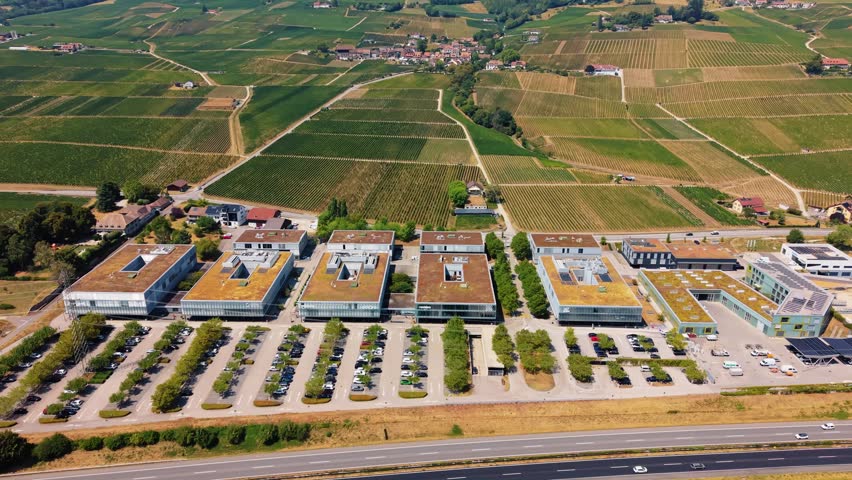 Aerial view of A-One Business Center with modern office buildings, green roofs, and parking lots surrounded by vineyards in Rolle, Vaud, Switzerland.