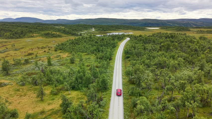 Aerial view of a red car driving on a road near Mittaklappen surrounded by lush green forests and mountains in Funasdalen, Jamtlands lan, Sweden.