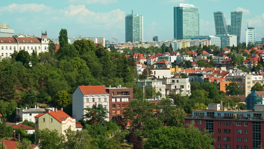 Historic residential houses lie surrounded by green trees in Prague. Modern glass skyscrapers rise in distant business district. Scenic urban view shows city contrast and real estate growth.
