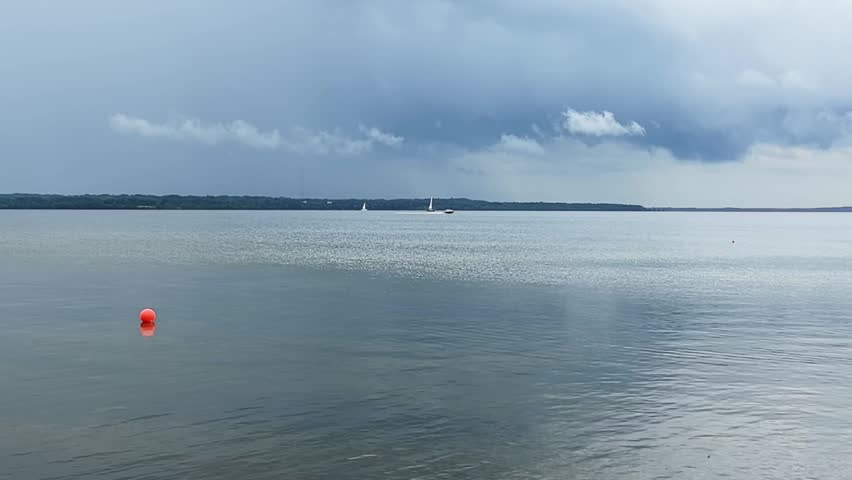 Wide view of a calm lake with distant sailboats and an orange buoy under dramatic gray storm clouds. Atmospheric nature b-roll footage.