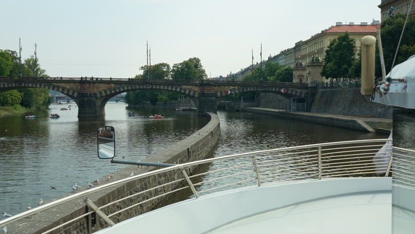 A tourist boat approaches Smichov Lock entrance on Vltava River in Prague, Czechia. Historic stone bridge spans across water. Czech waterway engineering shows European river cruise travel scene.