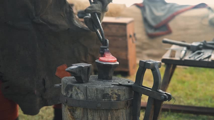 In a historical reenactment, a blacksmith works at an anvil, heating metal in a forge. The blacksmith wears traditional clothing, focusing on crafting tools in a rustic outdoor setting.