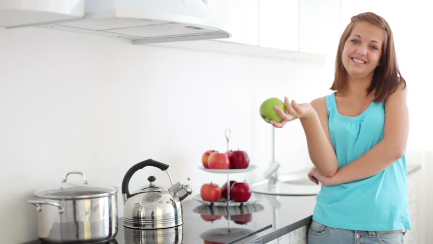 Girl standing in kitchen, holding green apple and laughing

