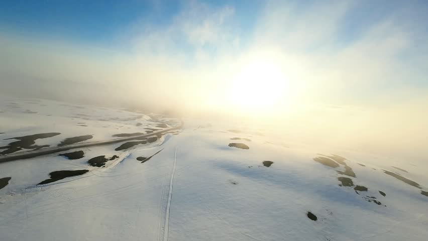 An aerial perspective of a vast snowy terrain dotted with forested islands and a visible trail or path cutting through the frozen landscape. The scene is bathed in soft, diffused light from a low sun creating a hazy, ethereal atmosphere typical of arctic or subarctic winter conditions.