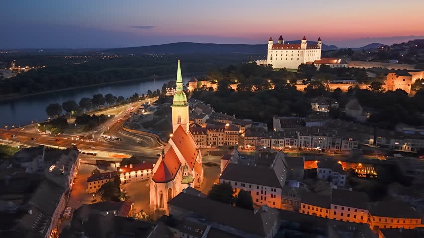 Bratislava castle and illuminated cathedral at dusk aerial view. Beautiful evening skyline over Bratislava old town and Danube river. Aerial view of historic Bratislava landmark buildings at sunset