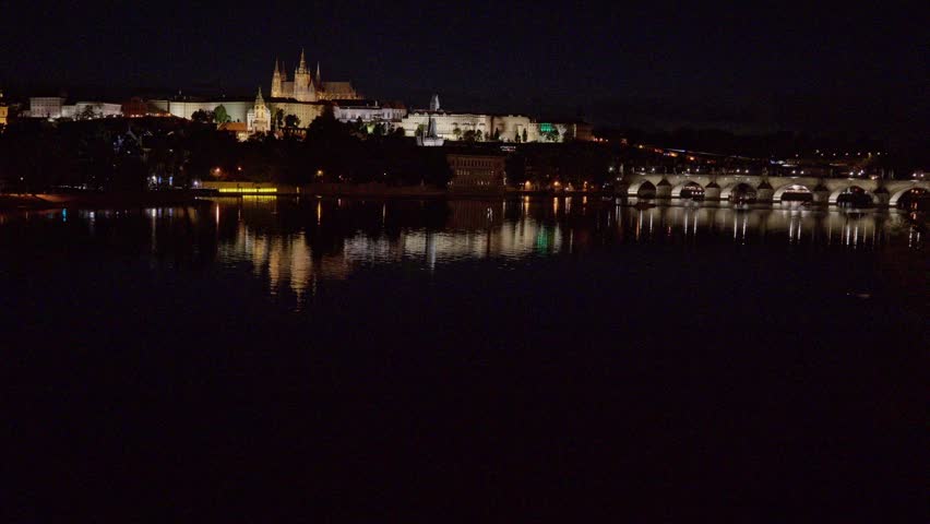 Wide shot of Prague Castle and illuminated Charles Bridge reflected in Vltava River at night, Czech Republic.