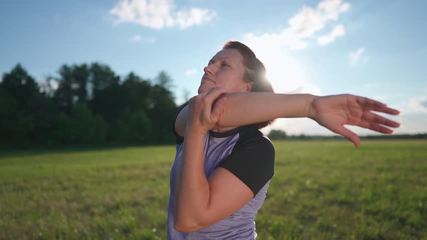 A woman is stretching in a grassy field as the sunlight reflects behind her. The woman is focused on her sport activity while enjoying the open space.