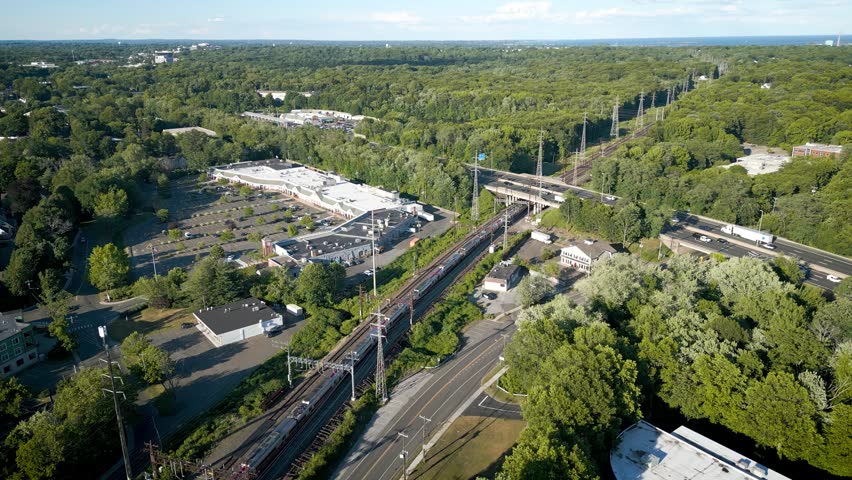 Aerial tracking shot of a commuter train leaving Darien Connecticut on railway tracks parallel to a busy interstate highway through a lush green suburban landscape.