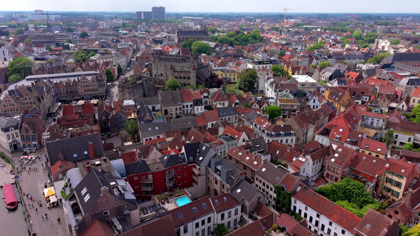 An aerial drone video shot providing a high-angle, panoramic overview of the historic city center of Ghent, Belgium. The video frame features a wide vista of dense residential neighborhoods packed with traditional Flemish gabled houses, terracotta red-tiled roofs, and modern urban infill. In the upper-left midground, the iconic Gravensteen medieval castle stands surrounded by its stone ramparts and canals. Narrow cobblestone streets, vibrant public plazas, and green courtyards weave through the tightly packed buildings, leading toward the distant skyline under an overcast daylight sky. The elevated drone angle captures the charming mix of historic preservation and dense European urban living. This high-definition video footage highlights Belgian architectural heritage, urban tourism, historical landscapes, and popular European travel destinations.