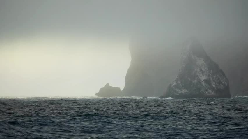 Atmospheric wide shot of dark, rough ocean waves rolling and splashing against massive sea cliffs shrouded in thick fog and mist on a stormy day.