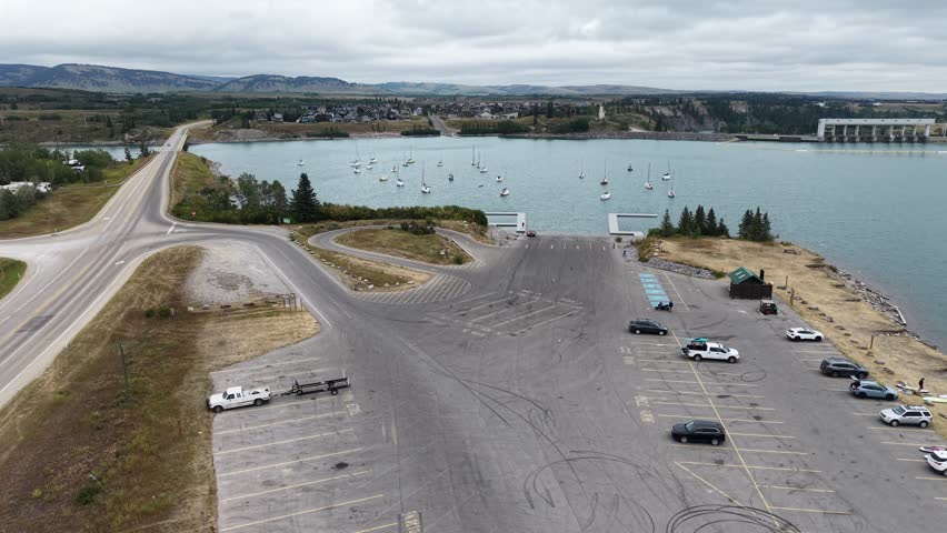 Aerial drone view of a lakeside parking area with several cars, a boat launch, and a calm lake in the background.