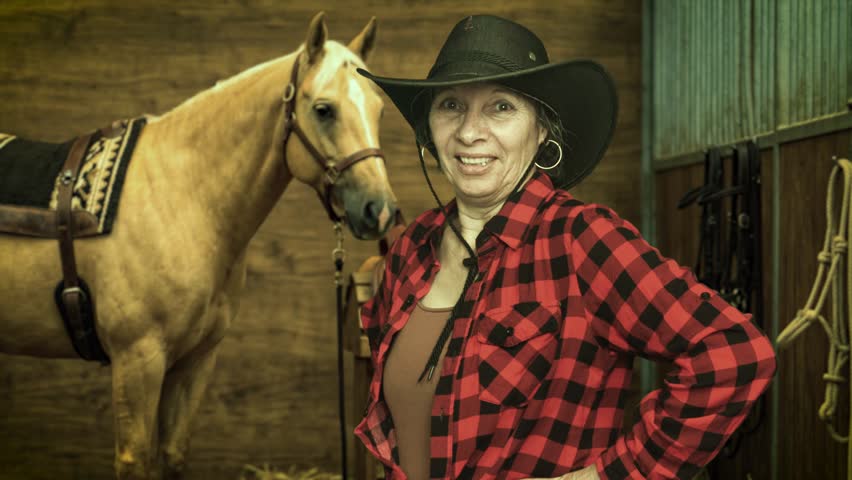 Smiling senior cowgirl tipping her hat beside a saddled palomino horse inside a stable. Western ranch lifestyle and equestrian portrait. 4K.