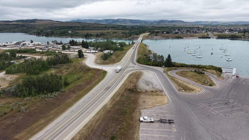 Aerial drone view of a lakeside road intersection with a parking area, boat launch, and calm lake surrounded by natural landscape.