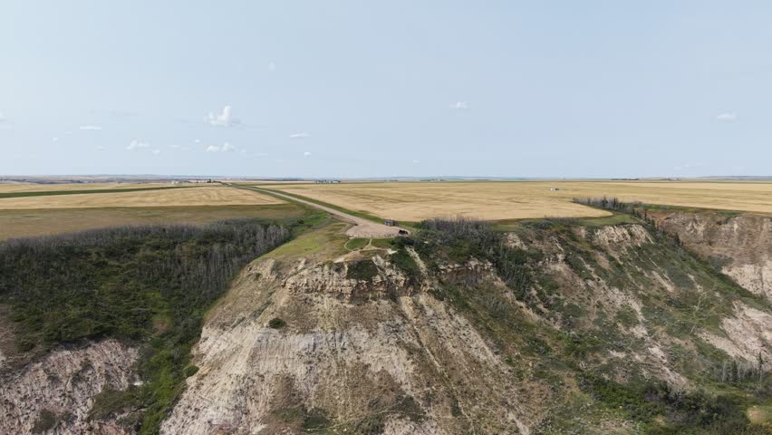 Aerial drone view of rugged prairie cliffs and open grassland, with exposed earth and agricultural fields stretching across the landscape.