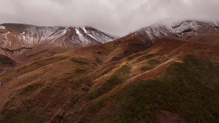 Twin snow streaked peaks rise beneath clouds above tussock foothills at Mount Ngauruhoe, Tongariro, New Zealand, rugged orange terrain slopes toward green covered ridges. Aerial drone wide shot