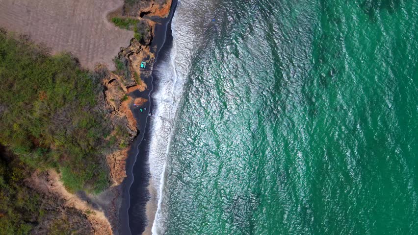 Turquoise waves roll against black sand and rocky cliffs at Playa Negra, with tropical vegetation lining the coast in Vieques, Puerto Rico, aerial drone descending