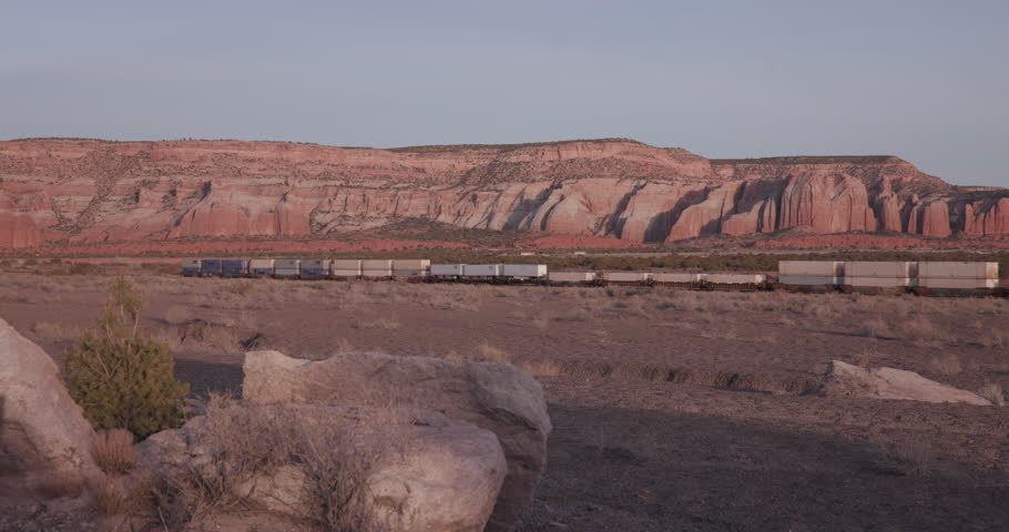 Cinematic footage of the tail end of a long freight train traveling through Gallup, New Mexico, around sunset. Intermodal shipping containers move along the railroad with the region’s distinctive red sandstone bluffs rising in the background, creating a classic American Southwest transportation scene. Ideal for freight, logistics, railroad, travel, industry, and Southwestern landscape productions.