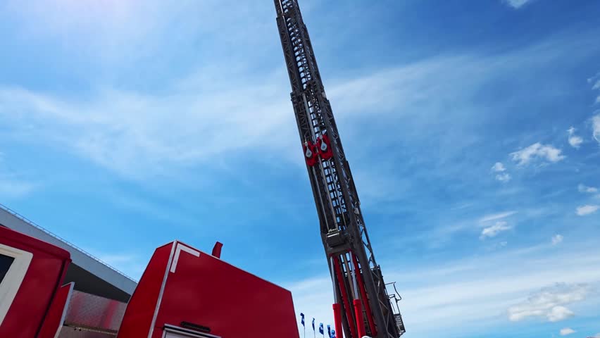 Extended ladder of fire truck reaching high into blue sky. Bright sun with lens flare. Wispy white clouds. Red truck body details, indicating emergency service.