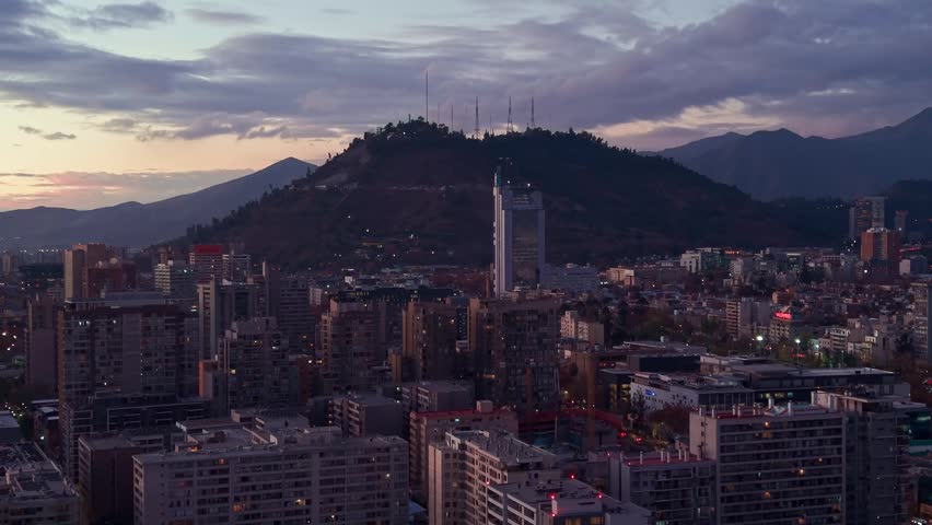 Approaching shot toward Cerro San Cristobal with antennas rising above a dense city neighborhood and mountains at dusk. Santiago, Chile