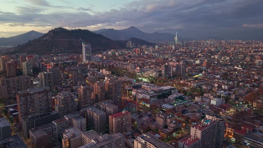 Forward flight tilting up over dense apartment rooftops revealing Cerro San Cristobal hill and distant skyline at dusk. Santiago, Chile