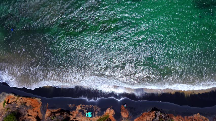 Aerial view of waves washing onto black sand beach with rocky shoreline and turquoise water