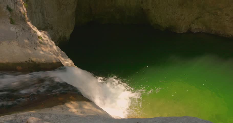 Small and charming water cascades over smooth rock into an emerald green watered pool surrounded by cliffs in bright daylight with gentle movement on the water surface.