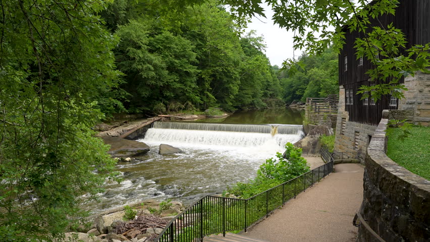 Static wide shot of a historic wooden mill building beside a river dam and waterfall surrounded by lush forest. Water cascades steadily over the stone spillway into a rocky stream below. A stone walkw