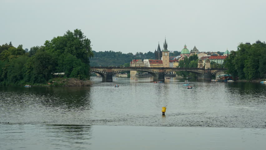 People ride pedal boats on Vltava River in Prague. Historic Charles Bridge stands in background. Scenic urban panorama depicts summer travel, European vacation, and water leisure activity.