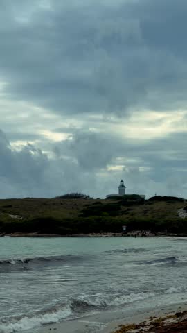 View of crashing ocean waves on a sandy beach shore near a historic white lighthouse on a hill in Puerto Rico