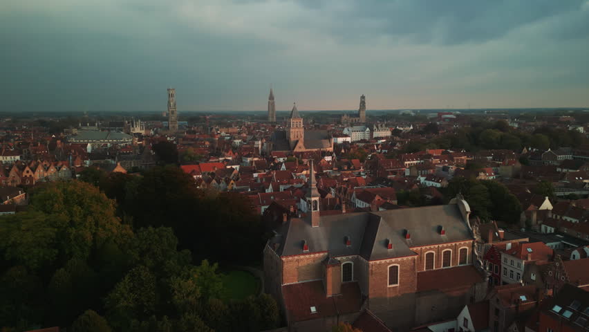 Historic church stands above autumn trees and red rooftops in Bruges, Belgium, while distant towers define the layered medieval skyline beneath soft morning clouds.