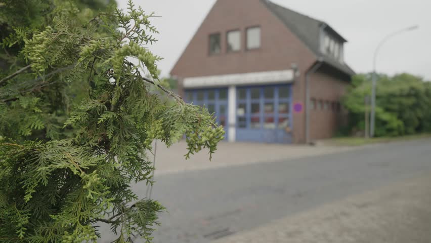 A wide shot captures a traditional small-town fire station building made of red brick, featuring blue garage doors housing a fire truck inside, surrounded by lush green foliage under an overcast sky.