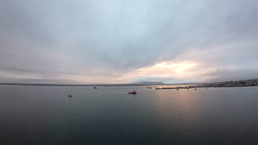 Harbor of Puerto Natales, Chile, with anchored vessels on calm water and a soft glow breaking through clouds over Última Esperanza Fjord, FPV drone footage