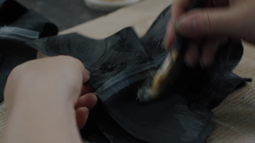 Hands applying adhesive to black shoe uppers with a brush in a workshop setting